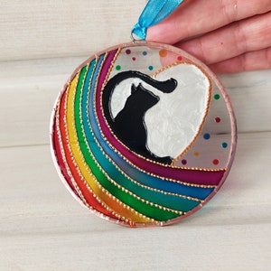 May include: A round glass ornament with a rainbow design and a black cat silhouette inside a white heart. The ornament is decorated with gold glitter and small colorful dots.
