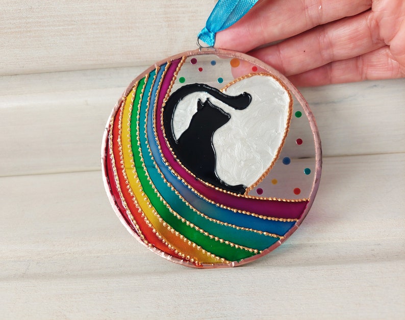 May include: A round glass ornament with a rainbow design and a black cat silhouette inside a white heart. The ornament is decorated with gold glitter and small colorful dots.