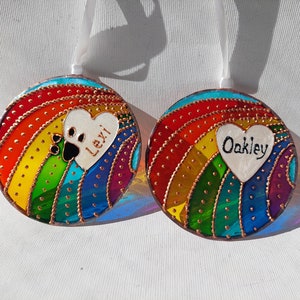 May include: Two round glass ornaments with a rainbow design and a white heart with a paw print and a name. The ornaments are decorated with gold glitter and have a white ribbon for hanging.