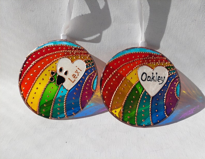 May include: Two round glass ornaments with a rainbow design and a white heart with a paw print and a name. The ornaments are decorated with gold glitter and have a white ribbon for hanging.