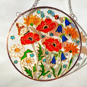 May include: A round, stained glass panel featuring a vibrant floral design. Red poppies, orange and yellow flowers, bluebells, and white daisies are depicted with green leaves. A butterfly and ladybug are also included. The panel is framed with a copper-colored border and has a chain for hanging.
