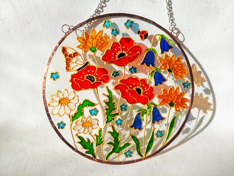 May include: A round, stained glass panel featuring a vibrant floral design. Red poppies, orange and yellow flowers, bluebells, and white daisies are depicted with green leaves. A butterfly and ladybug are also included. The panel is framed with a copper-colored border and has a chain for hanging.