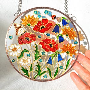 May include: A round, stained glass window featuring a floral design. The window showcases red poppies, white daisies, orange and yellow flowers, and blue bellflowers. A butterfly and ladybug are also included in the design. The window is framed with a copper-colored border and hangs from a chain.