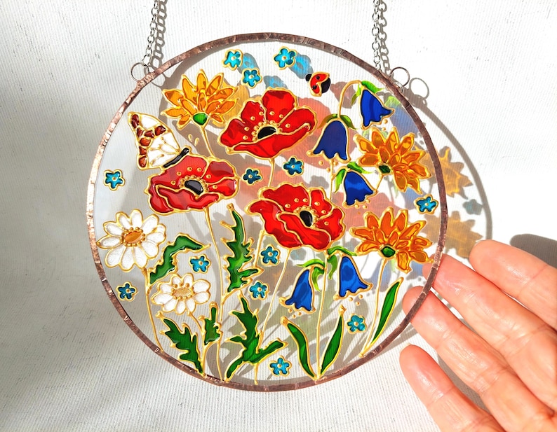 May include: A round, stained glass window featuring a floral design. The window showcases red poppies, white daisies, orange and yellow flowers, and blue bellflowers. A butterfly and ladybug are also included in the design. The window is framed with a copper-colored border and hangs from a chain.