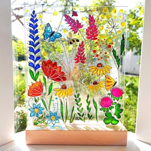 以下が含まれることがあります： 花、蝶、蜂が描かれた、カラフルなガラスパネル。花は赤、オレンジ、黄色、青で、緑の葉と茎があります。パネルは木製の台座に取り付けられています。
