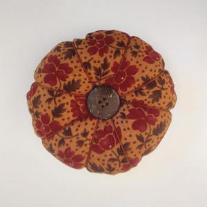 May include: A round, brown pincushion with a floral pattern in shades of red and brown. The pincushion has a button in the centre made of natural materials.