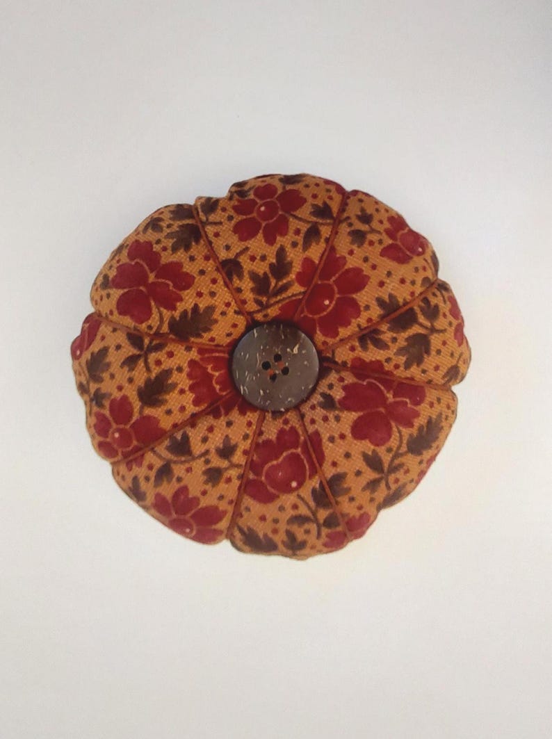 May include: A round, brown pincushion with a floral pattern in shades of red and brown. The pincushion has a button in the centre made of natural materials.