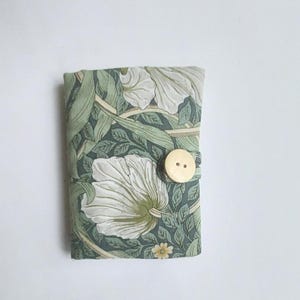 May include: A small, fabric-covered notebook or journal featuring a floral pattern in shades of green and white. The cover is secured with a gold-colored button. The design includes large white flower blooms and green foliage.