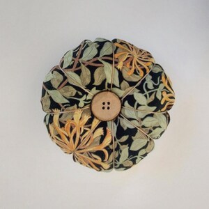 May include: A round pincushion with a floral pattern in shades of green, yellow, and black. The pincushion has a wooden button in the centre.