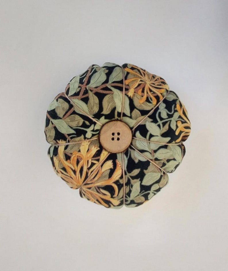 May include: A round pincushion with a floral pattern in shades of green, yellow, and black. The pincushion has a wooden button in the centre.