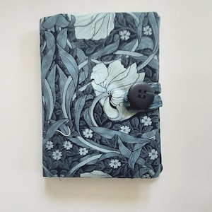 May include: A rectangular fabric-covered notebook featuring a dark blue and light blue floral pattern with long leaves and small blossoms. The cover is secured by a fabric tab with a single large, round, dark gray button centered on the right side.