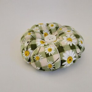 May include: A green and white floral pincushion with a white button in the centre. The pincushion is shaped like a flower and has a checkered pattern.