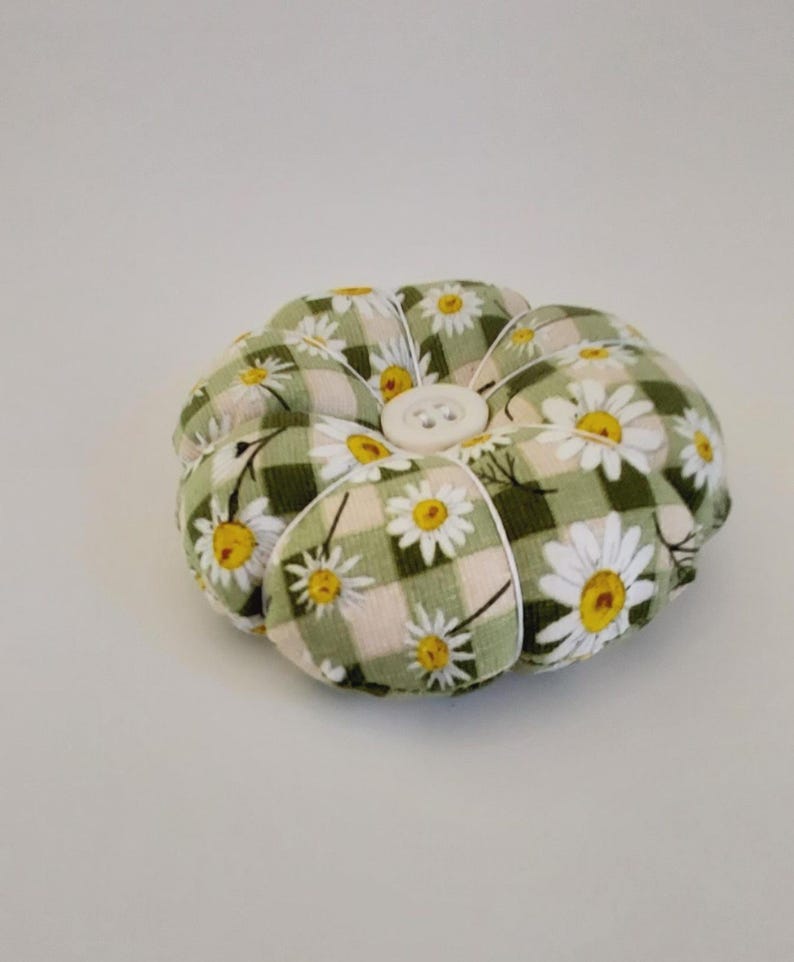 May include: A green and white floral pincushion with a white button in the centre. The pincushion is shaped like a flower and has a checkered pattern.