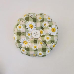 May include: A green and white gingham pincushion with a daisy print and a white button in the centre.