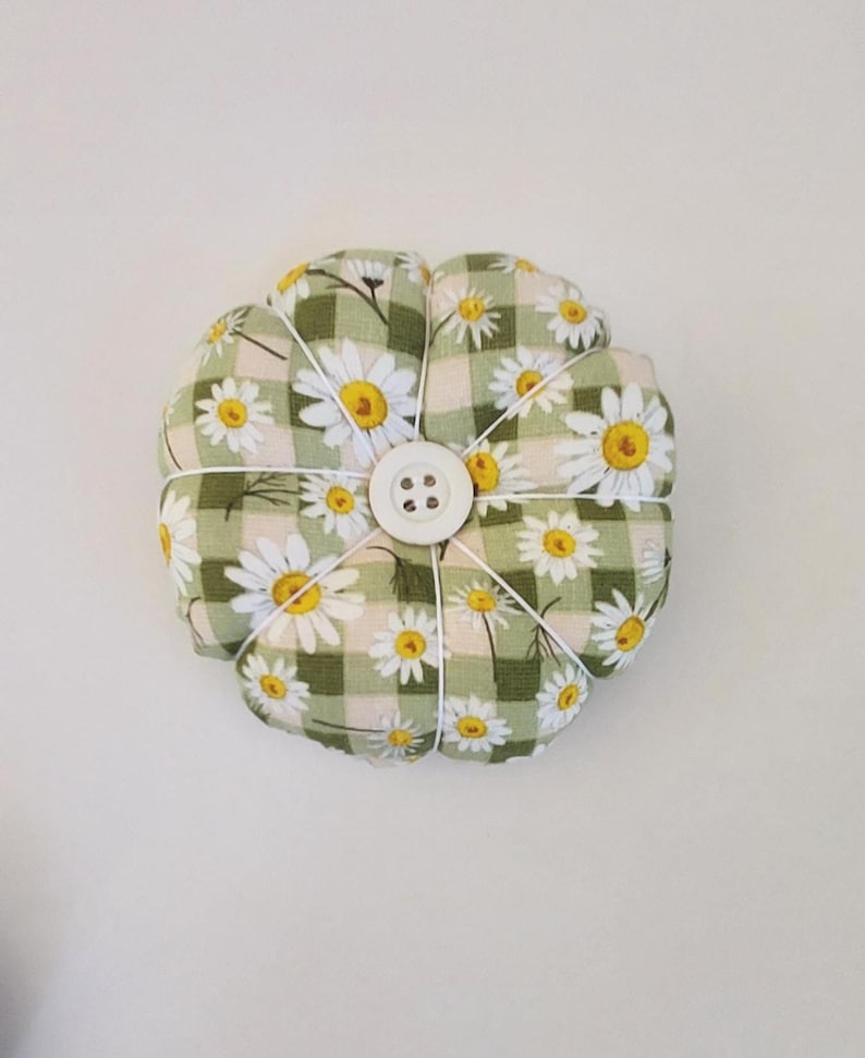 May include: A green and white gingham pincushion with a daisy print and a white button in the centre.