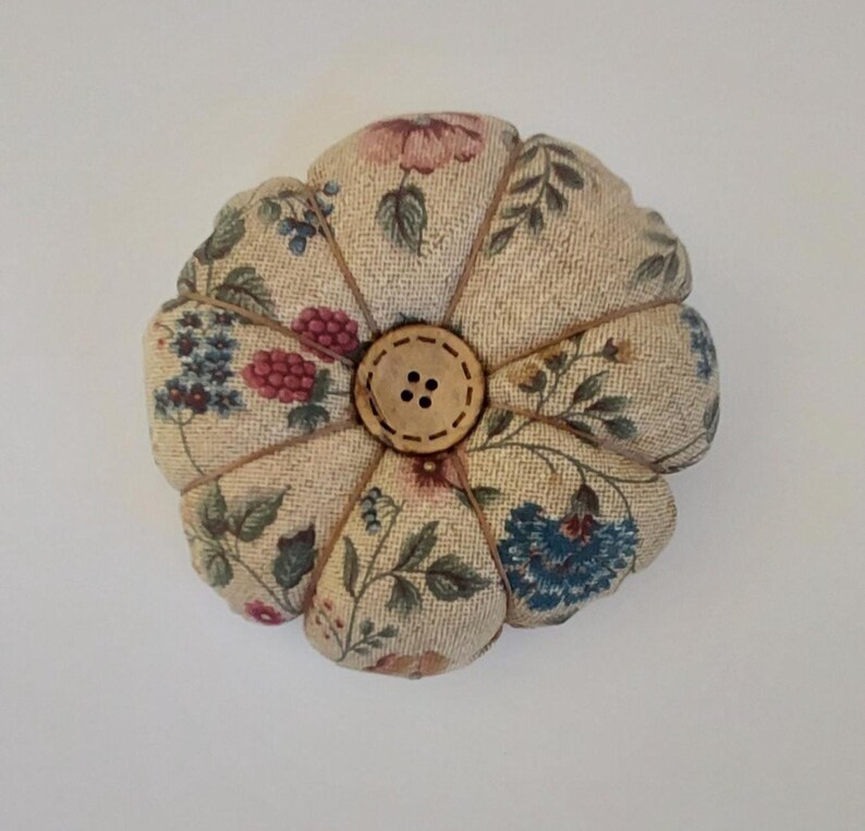 May include: A beige pincushion with a floral pattern and a wooden button in the centre. The pincushion is shaped like a flower with multiple petals.