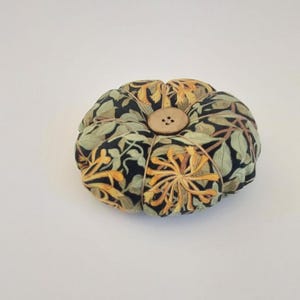 May include: A round pincushion with a floral pattern in shades of green, yellow, and black. The pincushion has a button in the centre.