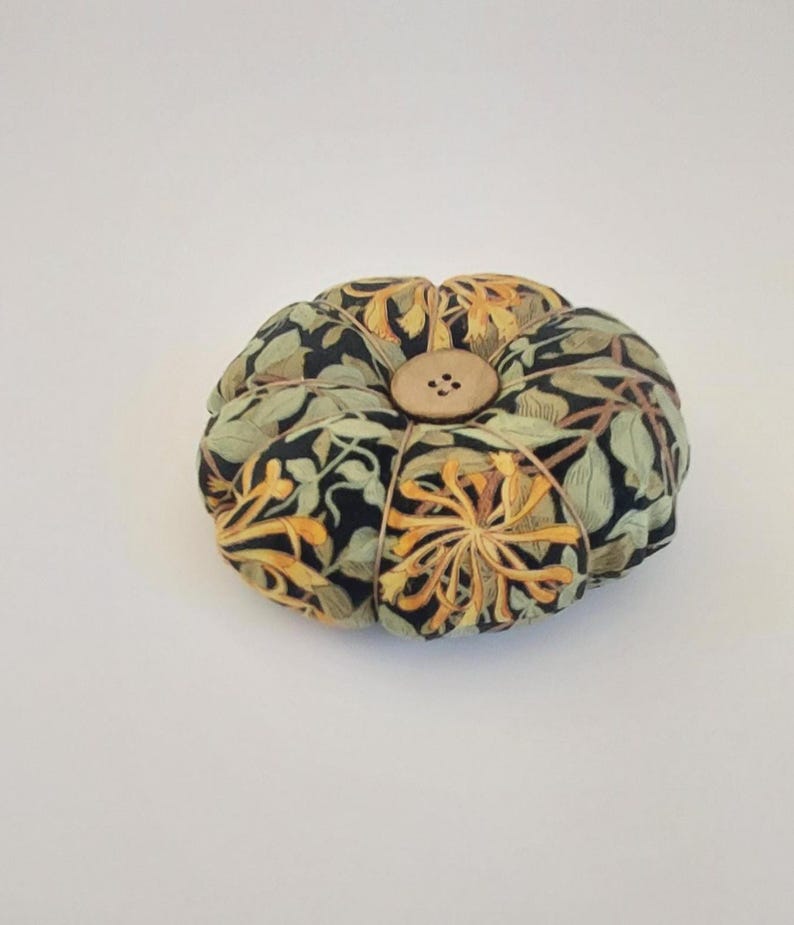 May include: A round pincushion with a floral pattern in shades of green, yellow, and black. The pincushion has a button in the centre.