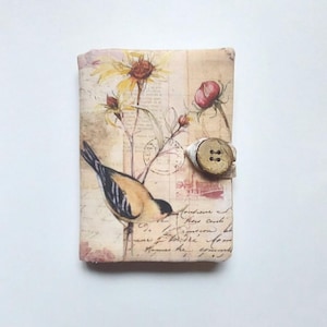 May include: A small, cream-colored fabric journal with a vintage floral and bird design. The journal has a button closure and a handwritten inscription on the front.