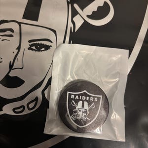 May include: A black and silver Raiders logo phone grip. The grip is round and features the Raiders shield logo with the team name and a helmeted player. The background includes a black and white graphic of a woman's face and a sword.