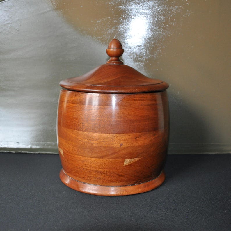May include: A wooden barrel-shaped container with a lid and a knob on top. The container is made of light brown wood with a darker brown wood trim. The container is sitting on a dark brown surface.