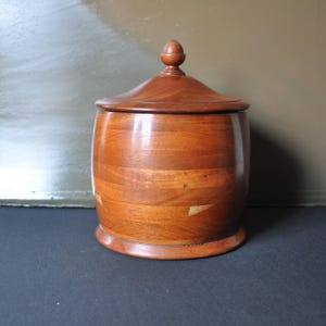 May include: A wooden container with a lid and a rounded shape. The container is made of light brown wood with a smooth finish. The lid has a small knob on top.