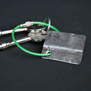 May include: Two silver keys attached to a green cord with a metal tag. The tag is rectangular and has a slightly textured surface.