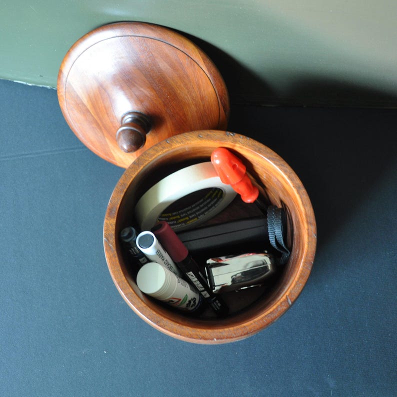 May include: A wooden container with a lid, open to reveal various items inside, including a roll of tape, a red screwdriver, a black marker, a white marker, a white tube, and a black and silver object.