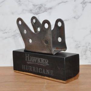 May include: A metal bracket with multiple holes sits atop a dark wooden block. The block is engraved with the words "Hawker Hurricane" in a vintage font. The bracket is a silver color and the wood is black.