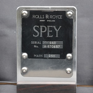 May include: A silver metal plaque with black lettering that reads "ROLLS R ROYCE DERBY ENGLAND SPEY SERIAL No. 660 A-670657 MARK 250".
