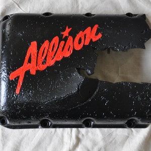 May include: A black metal transmission pan with a red "Allison" logo and a star. The pan is cracked and broken.