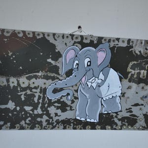 May include: A hand-painted elephant illustration on a weathered metal panel. The elephant is gray with pink ears and a white bib. The panel has a distressed, dark gray finish with visible rivets, creating a vintage aesthetic.