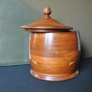May include: A wooden container with a lid. The container is made of light brown wood and has a rounded shape. The lid has a small knob on top. The container is sitting on a dark surface.
