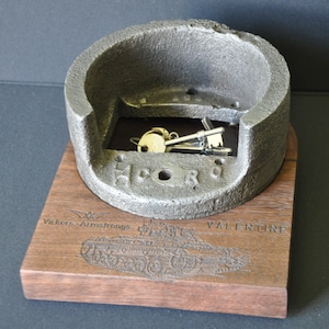May include: A gray metal key holder shaped like a tank turret, with the words "H.C.O.R.S" engraved on the side. The holder is mounted on a wooden base with an engraved image of a tank and the words "Vickers-Armstrongs" and "VALENTINE".