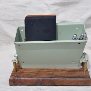 May include: A desk organizer made from a repurposed metal container, painted in a light green color. It features a dark blue wallet, pens, and pencils. The base is a dark wood with the text "Hawkon ScallHawk".