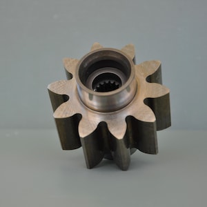 May include: A close-up of a metal gear with eight pointed teeth. The gear has a central cylindrical hub with a smaller toothed inner ring. The metal has a brushed finish, and the gear is set against a gray background.