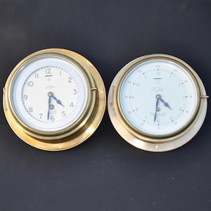May include: Two brass ship clocks with white faces and black hands. The clock on the left has Roman numerals and the clock on the right has Arabic numerals. Both clocks have a brass bezel.