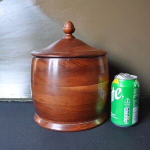 May include: A wooden barrel-shaped container with a lid and a knob on top. The container is made of dark wood and has a smooth finish. It is about the size of a standard soda can.