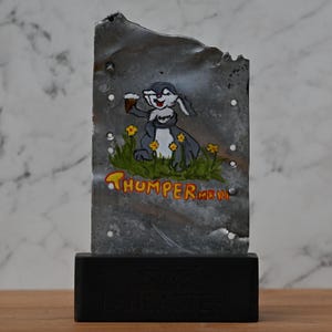May include: A metal artwork depicting a cartoon rabbit holding a drink, with the word "THUMPER" beneath. The rabbit is set in a grassy area with yellow flowers. The art is mounted on a black base with the word "LANCASTER" on it.