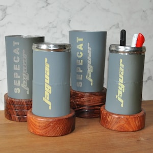 May include: Four cylindrical gray metal containers with silver rims, each mounted on a circular dark wood base. The containers feature the text SEPECAT jaguar printed in yellow. One container holds a black pen, a white pen, and a red pen.