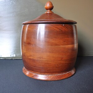May include: A wooden storage jar with a lid. The jar is a dark brown color and has a rounded shape. The lid has a small knob on top.
