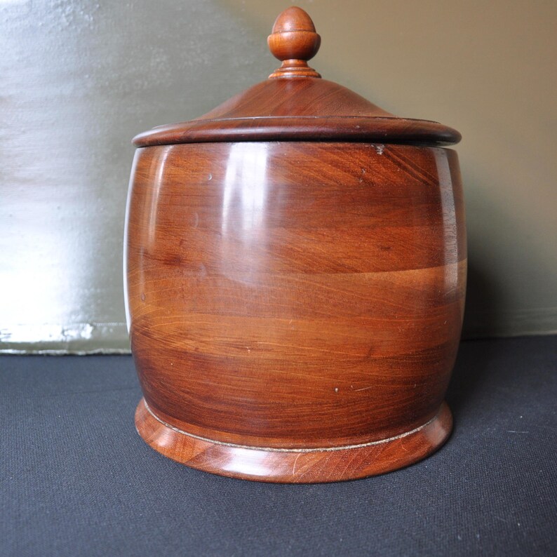 May include: A wooden storage jar with a lid. The jar is a dark brown color and has a rounded shape. The lid has a small knob on top.