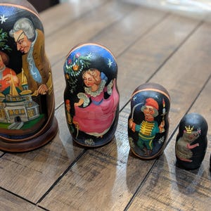 May include: A set of five Russian nesting dolls, each painted with detailed figures and scenes. The largest doll depicts a man and two children, while the others feature a woman, a soldier, a mouse, and a small floral design. The dolls are arranged on a wooden surface.