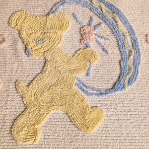 Op de afbeelding: Crèmekleurig textiel met een geel teddybeerontwerp en blauwe accenten. De beer houdt een blauwe en gele boog vast met een roze ster. Roze en blauwe accenten zijn verspreid over het textiel.