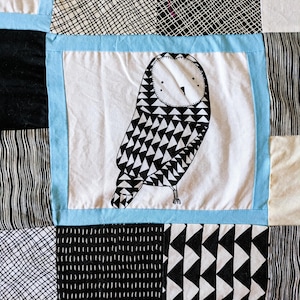 May include: A patchwork quilt featuring a central square with a black and white owl illustration. The quilt incorporates various black and white patterned squares, including geometric and linear designs, framed by a light blue border.