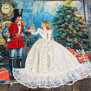 May include: A square decorative pillow cover featuring a printed scene of a nutcracker in a red uniform and a figure in a white lace dress standing before a Christmas tree with red ornaments and gifts, accented with a fabric flower and lace trim.