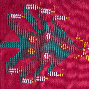 May include: Close-up of a red textile with an embroidered design. The design features a central geometric shape in green, white, and yellow. The background is a solid, textured red fabric, showcasing the detailed craftsmanship.