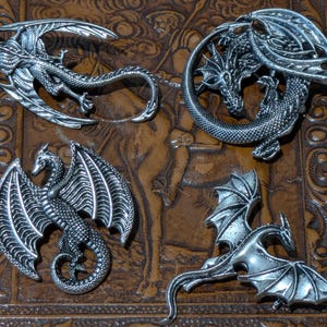 Puede incluir: Cuatro broches de dragón de metal plateado con detalles de escamas intrincados y alas extendidas. Cada broche representa un dragón en una pose diferente, incluyendo uno enroscado en círculo, uno volando con las alas abiertas y dos de pie.