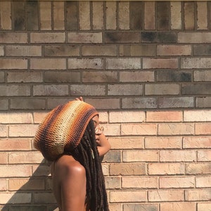 Puede incluir: Una persona con un gorro de ganchillo marrón, naranja y blanco con rastas largas. El gorro es de estilo con capucha y la persona está de pie frente a una pared de ladrillo.