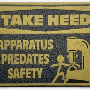 May include: A black metal sign with gold lettering that reads "Take Heed Apparatus Predates Safety". The sign features a silhouette of a person operating a machine.